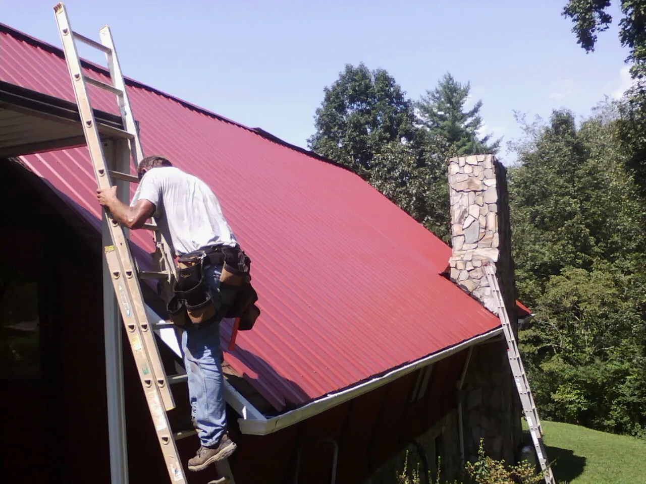 Roofer on a ladder beside a red metal roof and stone chimney — Soffit & Fascia Repair by Roofing Brothers in Melcroft